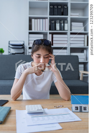 Young woman analyzing financial documents at home with calculator and model house on table in modern living room Young woman analyzing financial documents at home with calculator and model house on table in modern living room 123299709