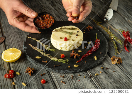 Hands skillfully sprinkle vibrant spices over a round cheese wheel, surrounded by fresh herbs, lemon, and garnishes. The rustic wooden table enhances the culinary artistry and warmth of the moment 123300081