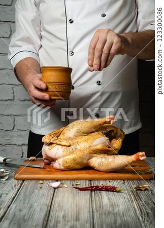 A passionate chef sprinkles salt on a whole chicken placed on a wooden cutting board. In the background, a rustic brick wall and cooking tools create a cozy atmosphere for cooking 123300086