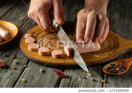 Hands skillfully slice sausage on a wooden cutting board. Nearby, a small dish holds various spices, adding flavor to the meal preparation process 123300098