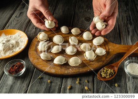 Dumplings are being arranged on a wooden board, surrounded by bowls of flour and spices. This culinary activity takes place on a rustic table, showcasing traditional cooking methods 123300099