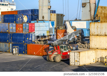 Two reach stackers and cranes transporting large cargo containers at shipping port. Bright sunlight, clear blue sky, stacked containers of various colors, shipping operations of sea cargo port. Two reach stackers and cranes transporting large cargo containers at shipping port. Bright sunlight, clear blue sky, stacked containers of various colors, shipping operations of sea cargo port. 123301536
