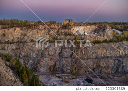 A rocky cliff with sparse trees and dry bushes under warm sunlight. At the top, an abandoned industrial structure stands among autumn foliage, creating a contrast between nature and human-made ruins. 123301889