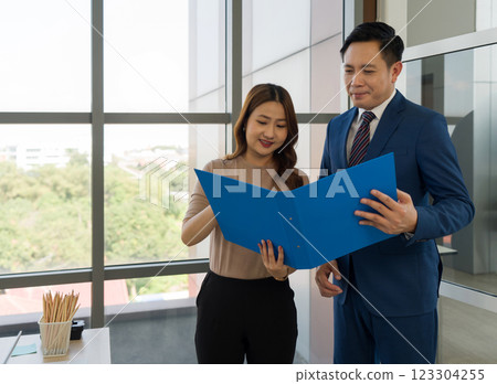 A collaborative work effort, a discussion on business report. Young secretary and manager in suit looking at document inside a blue folder. They are standing in a well-lit office with a large window 123304255