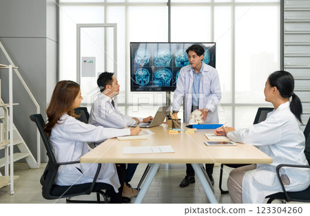 Young medical professional standing and presenting, holding a model of human skull and referencing image of brain scan displayed on a screen. The audience listening attentively with the presentation. Young medical professional standing and presenting, holding a model of human skull and referencing image of brain scan displayed on a screen. The audience listening attentively with the presentation. 123304260