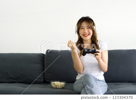 Young asian woman waring comfortable cloth and headphone, play console game in the living room. A glass bowl full of popcorn are on gray sofa. Young asian woman waring comfortable cloth and headphone, play console game in the living room. A glass bowl full of popcorn are on gray sofa. 123304370