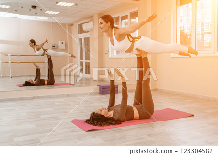 Two women do acroyoga on a light floor near a large mirror in a yoga studio. They are wearing leggings and tops. 123304582