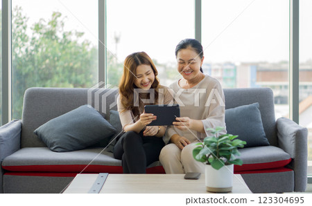 Two women are sitting on a couch, laughing together and sharing a tablet computer, in a bright living room. Two women are sitting on a couch, laughing together and sharing a tablet computer, in a bright living room. 123304695