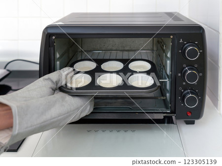 Hand with oven gloves, placing a muffin tray with batter into a black toaster oven in a white kitchen. 123305139