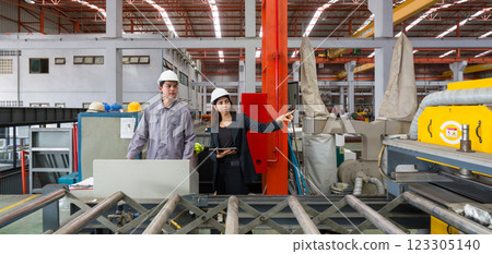 Two worker with safety helmet and black suit standing in front of metal sheet roofing machine, holding tablet computer used for taking note, checking schedule and reviewing digital document. Two worker with safety helmet and black suit standing in front of metal sheet roofing machine, holding tablet computer used for taking note, checking schedule and reviewing digital document. 123305140