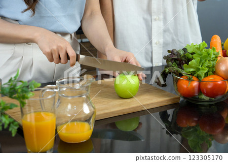 Closeup hand holding knife cutting green apple on a wooden chop board. Jar with mixed fruit juice and glass bowl with a variety of fruits and vegetables is placed on the kitchen counter. 123305170