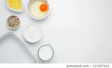 Flat lay composition for cooking preparation. Empty white baking dish with ingredients for baking, including egg, milk, butter, sugar, and nuts Flat lay composition for cooking preparation. Empty white baking dish with ingredients for baking, including egg, milk, butter, sugar, and nuts 123307422