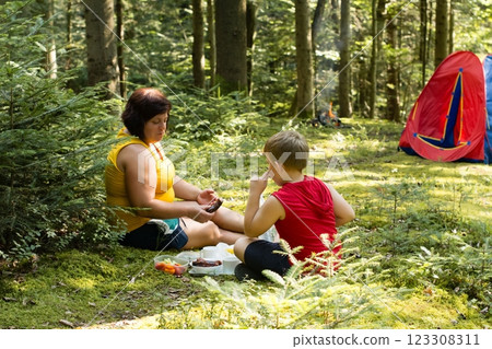A woman and child enjoy a picnic lunch in a forest setting, near a tent. 123308311