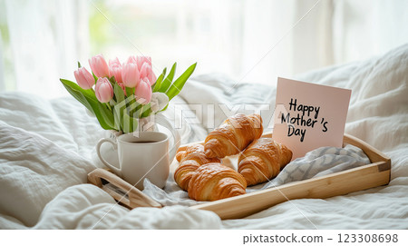 A cozy Mothers Day breakfast in bed with fresh croissants, coffee, pink tulips, and a handwritten card on a wooden tray. 123308698