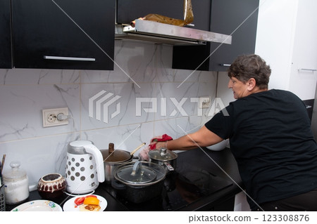 Woman cleaning a kitchen stove. She's wiping down the black countertop with a red cloth, surrounded by pots and a kettle on the stove. 123308876