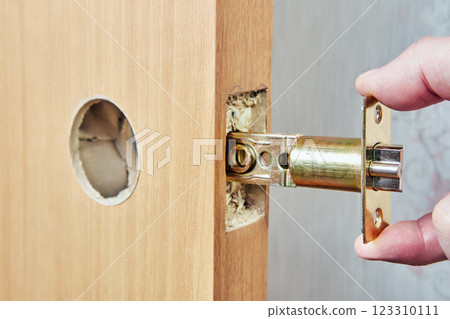 Latch bolt assembly, showcases brass strike plate being held against wood door edge, demonstrating installation with exposed mortise and circular knob hole. Latch bolt assembly, showcases brass strike plate being held against wood door edge, demonstrating installation with exposed mortise and circular knob hole. 123310111