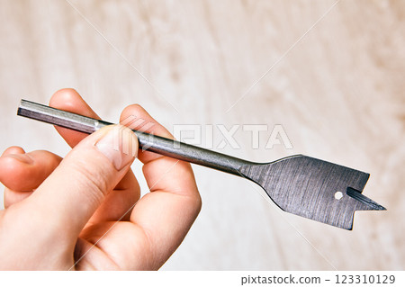 Hand holds metal spade bit used for drilling wide holes in wood, showcasing its flat blade with pointed spur for guiding and a shank for connecting to drill. Hand holds metal spade bit used for drilling wide holes in wood, showcasing its flat blade with pointed spur for guiding and a shank for connecting to drill. 123310129