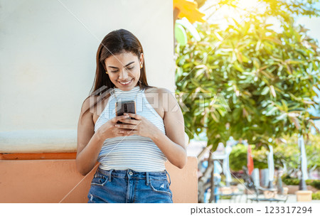 Happy girl with cell phone leaning against a wall. Young girl leaning against a wall messaging on her cell phone, Smiling young woman texting on cell phone outdoors Happy girl with cell phone leaning against a wall. Young girl leaning against a wall messaging on her cell phone, Smiling young woman texting on cell phone outdoors 123317294