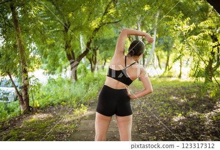 Athlete girl doing warm-up exercises in a park. Sporty woman doing stretching exercises before running. Athlete girl stretching arms before running. Concept of physical activity and healthy life 123317312