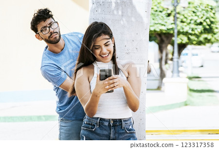 Jealous boyfriend spying on his girlfriend's cell phone in the park, Jealous man spying on his girlfriend texting on the cell phone. Suspicious man spying on his girlfriend with his cell phone 123317358