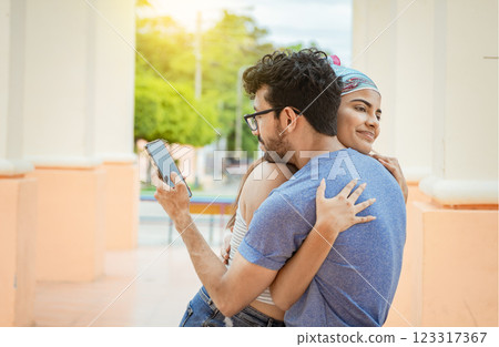 Unfaithful boyfriend hugging his girlfriend and looking at the phone. Unfaithful man looking cell phone while hugging his girlfriend outdoors, Concept of unfaithful man using cell phone Unfaithful boyfriend hugging his girlfriend and looking at the phone. Unfaithful man looking cell phone while hugging his girlfriend outdoors, Concept of unfaithful man using cell phone 123317367