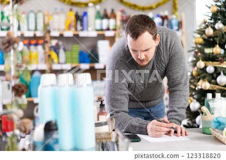 Attentive male salesperson restock of goods, checking papers in small household store decorated for New Year celebration Attentive male salesperson restock of goods, checking papers in small household store decorated for New Year celebration 123318820