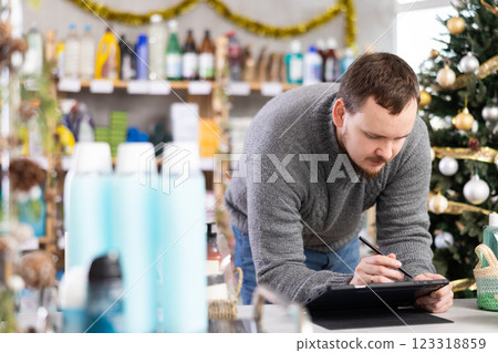 Bearded man seller restock of goods using tablet in store decorated for New Year holiday Bearded man seller restock of goods using tablet in store decorated for New Year holiday 123318859