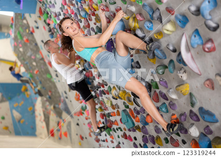 Adult woman is mastering climbing on training wall in gym, side view. Young woman holds on tightly to ledges and strives for top of bouldering route 123319244