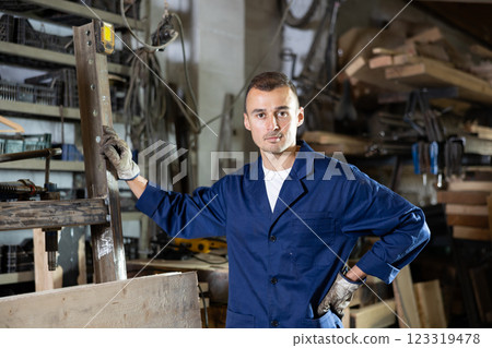 Male employee of workshop stands next to iron rails of workpieces, measures length 123319478