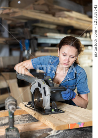 Working at sawmill - woman saws wooden board using circular saw Working at sawmill - woman saws wooden board using circular saw 123319542