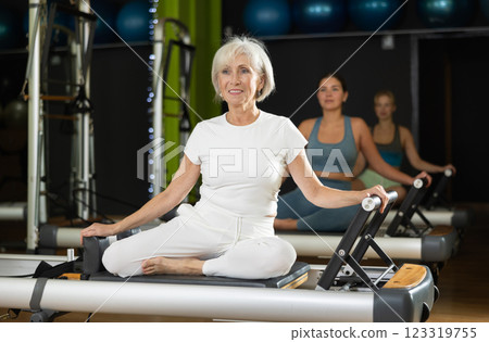 Mature woman sitting on foldable bed reformer performs exercises, strengthening body and muscles Mature woman sitting on foldable bed reformer performs exercises, strengthening body and muscles 123319755