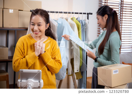 Two young asian woman live online clothing store while showing clothing with tablet and laptop. 123320780