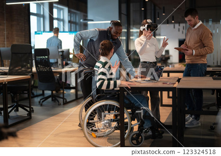 Disabled businesswoman in a wheelchair working in modern coworking office. Team around is using virtual reality goggles, collaborating with the latest technology in an open space startup creative 123324518