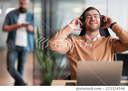 Excited businessman wearing headphones, sitting at his desk in a modern open-space startup office. Overjoyed executive enjoying his break, singing and listening to favorite music, having fun at work. Excited businessman wearing headphones, sitting at his desk in a modern open-space startup office. Overjoyed executive enjoying his break, singing and listening to favorite music, having fun at work. 123324546