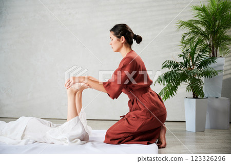 Masseuse making thai yoga massage. Woman therapist doing traditional massage treatment, stretching female patient's body. Client lying on mat, enjoying hand massage in calm atmosphere. 123326296