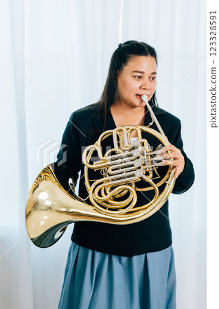 A woman stands at home in front of elegant curtains, playing a shiny gold horn. The warm lighting and her graceful demeanor bring out the beauty of her musical performance. 123328591