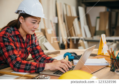 Carpenter woman in a white hardhat and red plaid shirt uses a laptop in a woodshop, surrounded by tools, wood scraps, and pencils. Ideal for online learning, client orders, or woodworking projects 123328596