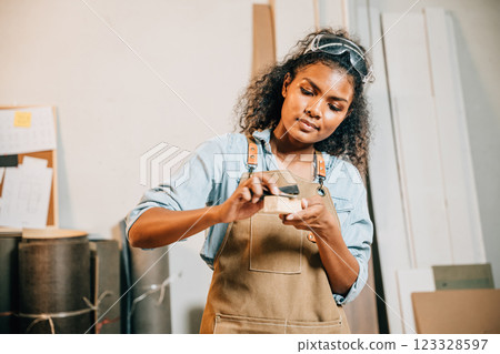 African American carpenter woman sanding a piece of wood. Wood grinding with sandpaper as she prepares timber for furniture making in the workshop, National Carpenters Day African American carpenter woman sanding a piece of wood. Wood grinding with sandpaper as she prepares timber for furniture making in the workshop, National Carpenters Day 123328597