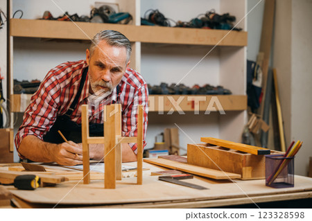 Senior carpenter in plaid shirt and overalls inspects a wooden stool in a workshop, surrounded by tools and materials. Workshop wall with tools visible in the background, showcasing craftsmanship Senior carpenter in plaid shirt and overalls inspects a wooden stool in a workshop, surrounded by tools and materials. Workshop wall with tools visible in the background, showcasing craftsmanship 123328598