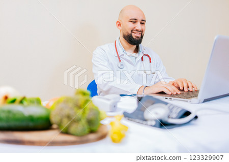 Smiling nutritionist with laptop at desk. Nutritionist doctor using laptop at workplace. Bearded nutritionist doctor working on laptop at desk. Smiling nutritionist with laptop at desk. Nutritionist doctor using laptop at workplace. Bearded nutritionist doctor working on laptop at desk. 123329907