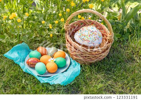 Easter cake in a wicker basket and a plate with colored Easter eggs on green grass with yellow Persian buttercup. Easter cake in a wicker basket and a plate with colored Easter eggs on green grass with yellow Persian buttercup. 123331191