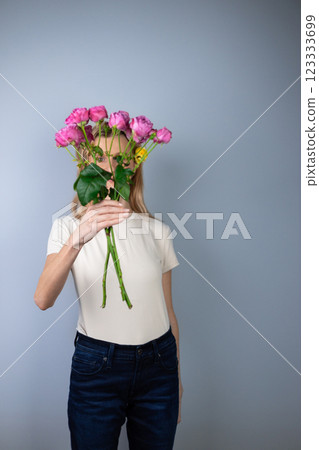 on light gray background a young blonde girl in a light T-shirt covers her face with pink peony roses March 8 on light gray background a young blonde girl in a light T-shirt covers her face with pink peony roses March 8 123333699