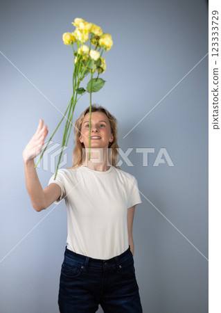 on light gray background a young blonde girl in a light T-shirt throws peony roses in front of her March 8 yellow flowers for separation smile on her face on light gray background a young blonde girl in a light T-shirt throws peony roses in front of her March 8 yellow flowers for separation smile on her face 123333729