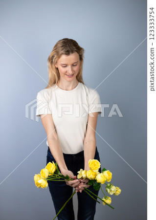 on light gray background a young blonde girl in a light T-shirt in her hands yellow peony roses March 8 bouquet on outstretched arms looking down 123333754