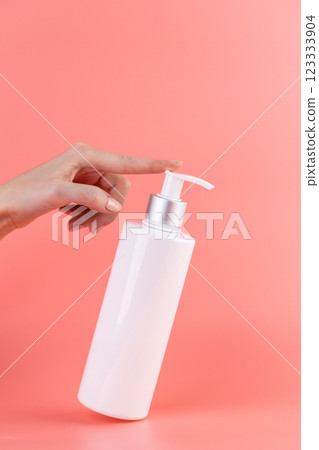 close-up on a soft pink background white glossy bottle with dispenser minimalist container for cosmetics beauty industry hermetic storage of liquids female hand presses the dispenser with a finger 123333904