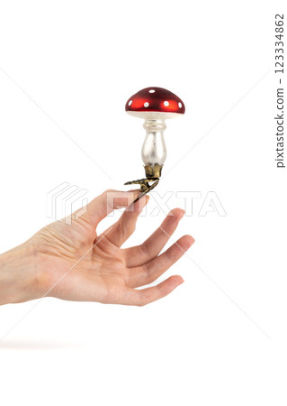 on white background, a woman's hand holds a glass enameled Christmas tree toy in the shape of an amanita mushroom with a clip at the base, modern decor toy with sparkles 123334862