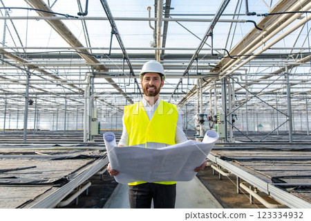 Young bearded engineer in hardhat working with sketches in a greenhouse structure Young bearded engineer in hardhat working with sketches in a greenhouse structure 123334932