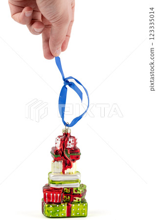 close-up on white background a man's hand holds a glass enameled Christmas tree toy in the shape of a stack of gifts in wrapping paper hanging blue ribbon modern decor toy with sparkles close-up on white background a man's hand holds a glass enameled Christmas tree toy in the shape of a stack of gifts in wrapping paper hanging blue ribbon modern decor toy with sparkles 123335014