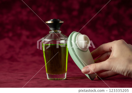 red burgundy velvet background there is a glass jar with green massage oil, on which a gently mint vacuum vibrator is leaning color device for girls relaxation for adults female hand takes a vibrator 123335205