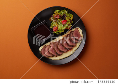 on orange background on a rectangular black plate lies mashed potatoes on which pieces of filet mignon are laid out, served with mushrooms and tomato on orange background on a rectangular black plate lies mashed potatoes on which pieces of filet mignon are laid out, served with mushrooms and tomato 123335330
